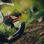 Lähikuva henkilöstä, joka ajaa Tunturi EMAX *SUOMEN EDULLISIN* sähköfatbike -pyörällä metsäpolulla, korostaen takapyörää, Shimano-keskimoottoria, vaihteita ja läskirengasta. Vihreät lehdet kehystävät kuvaa, vangiten liikkeen ja luonnon tuntua.