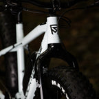 Lähikuva SÄHKÖPYÖRÄT.NETin Rock Machine Vyöry E50R:stä - valkoisesta e-fatbike-pyörästä, jossa on ohjaustanko, etuhaarukka, paksu Maxxis-rengas, geometrinen musta logo rungossa ja Shimano EP6 -moottori, joka parantaa suorituskykyä.